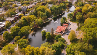 Aerial view of Washington Park’s lake lined with lush spring trees, with two small fountain sprays in the water, a red-roof lakehouse/boathouse on the shoreline, and a footbridge in the distance near surrounding neighborhoods.